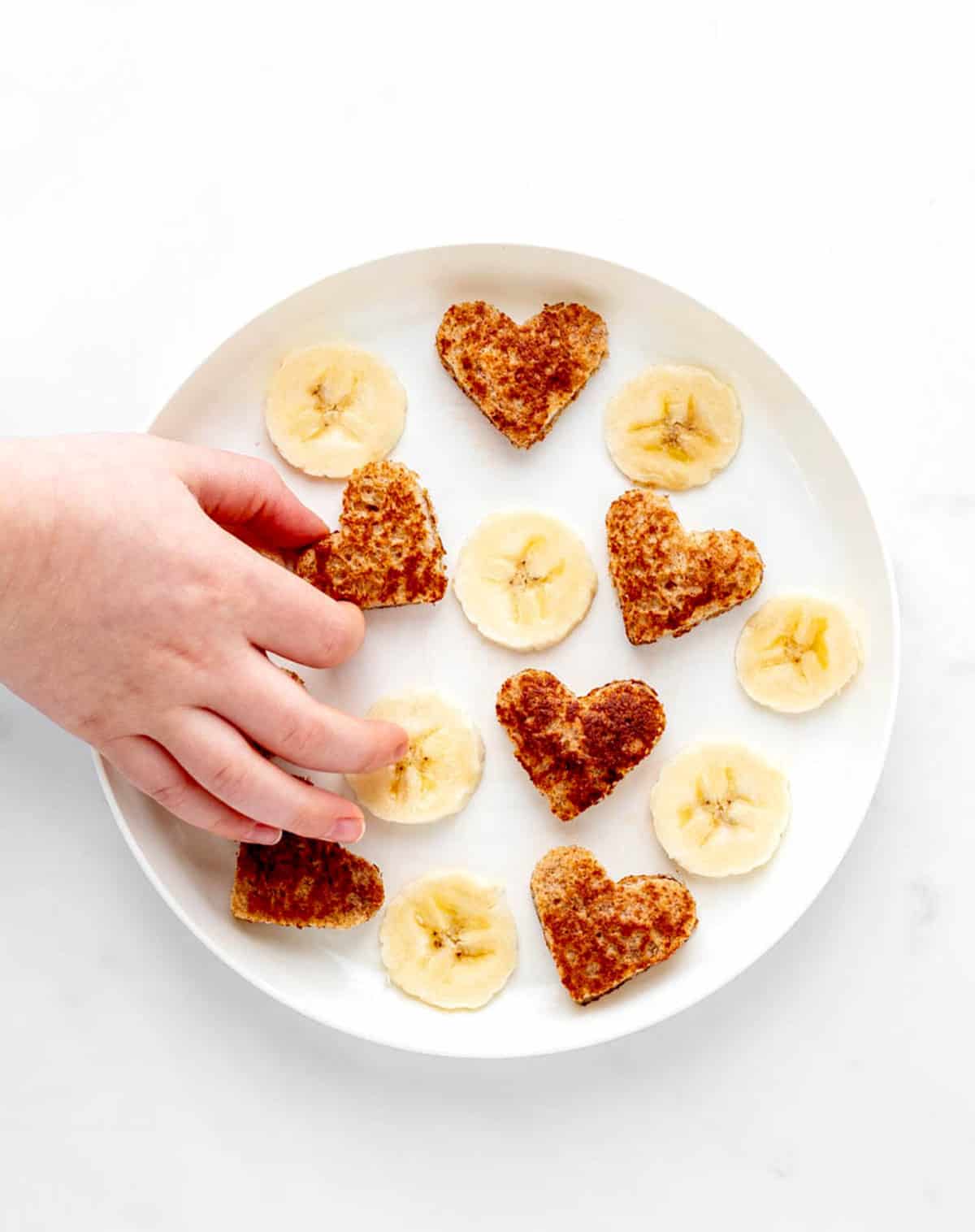 A white plate with banana French toast cut into heart shapes and banana slices with a child's hand picking up one of the toast hearts.