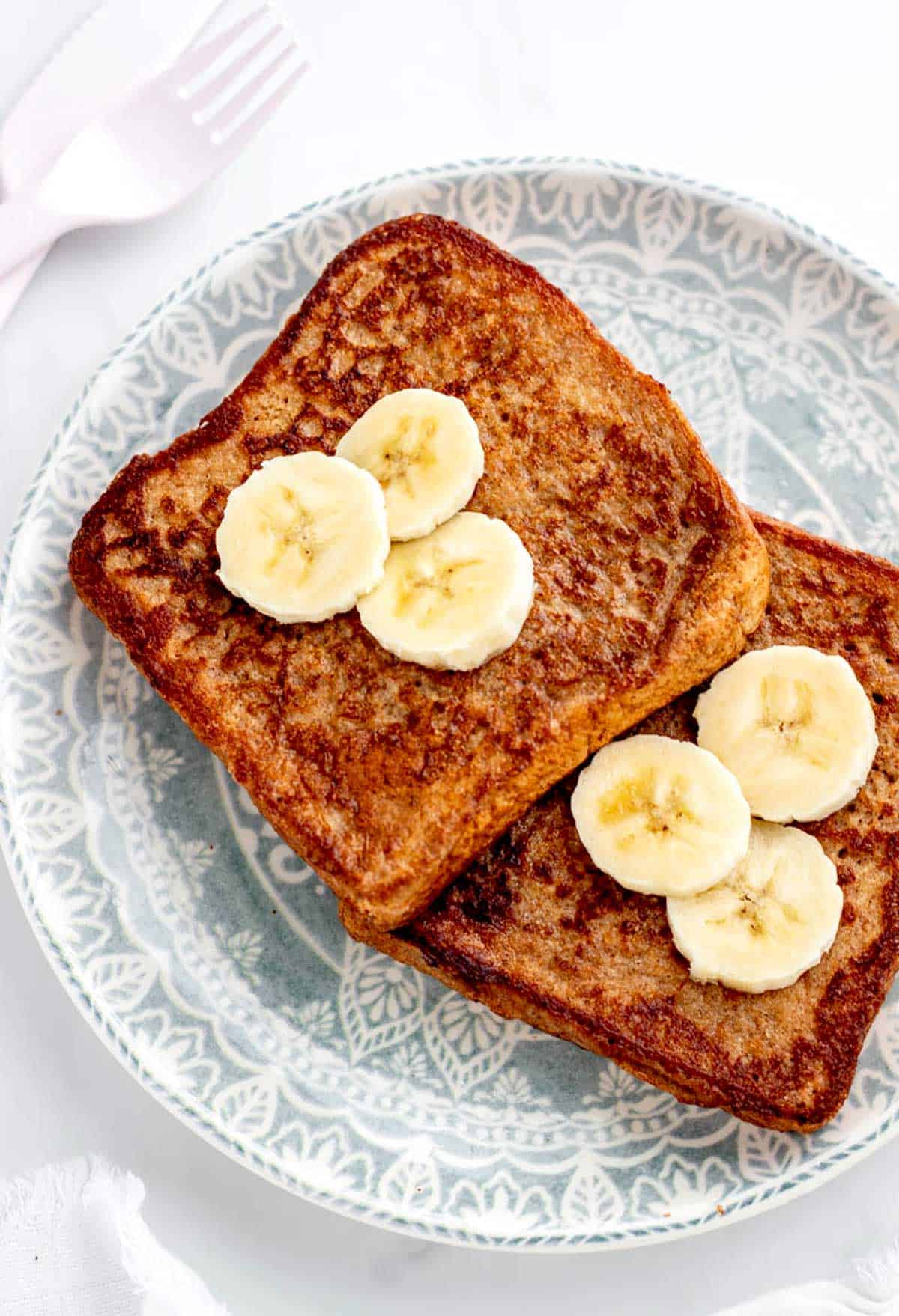 A close up image of two slices of banana French toast on a blue and white plate topped with slices of banana.