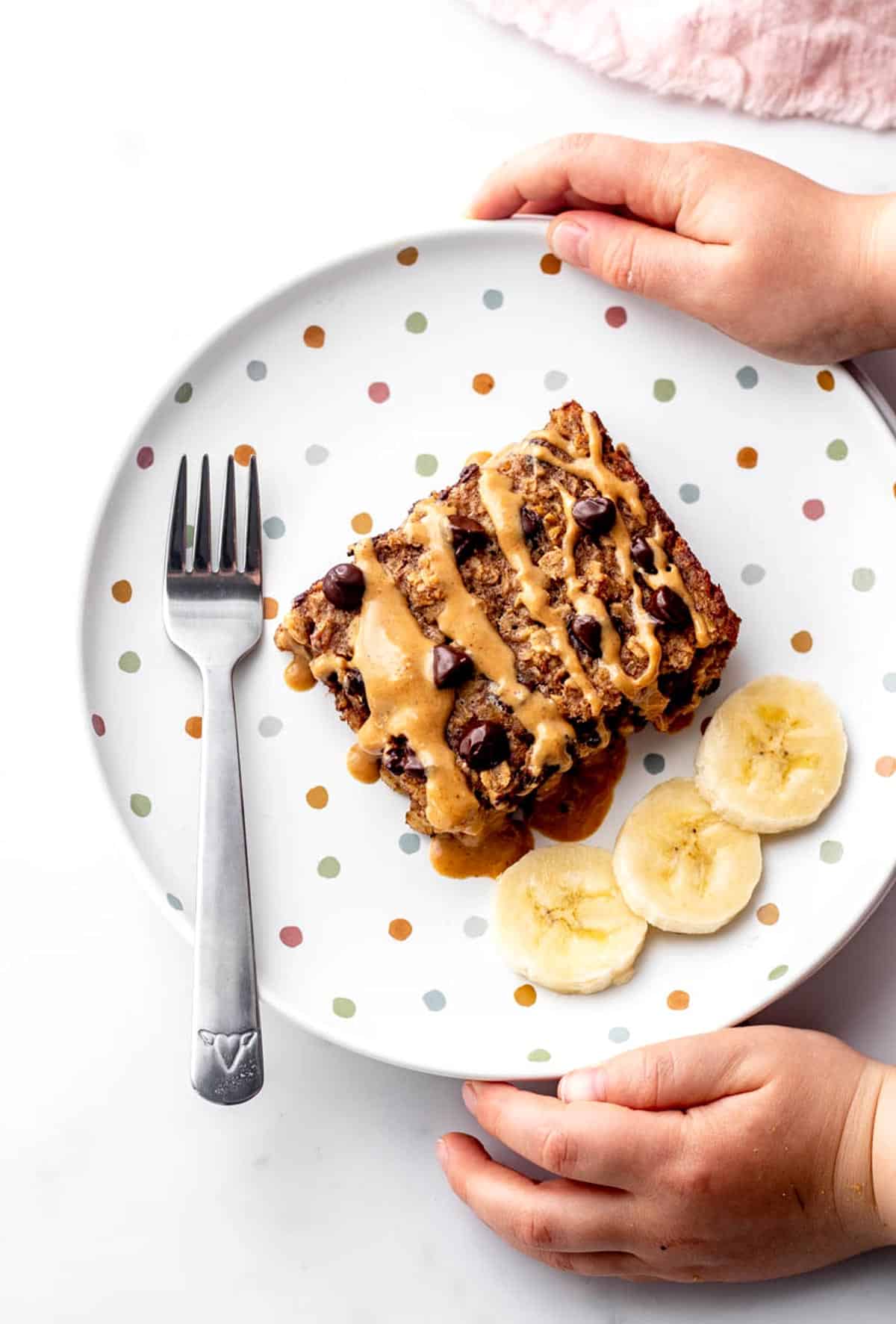 A child's hands cupping a plate with a slice of chocolate chip baked oats next to some banana slices.