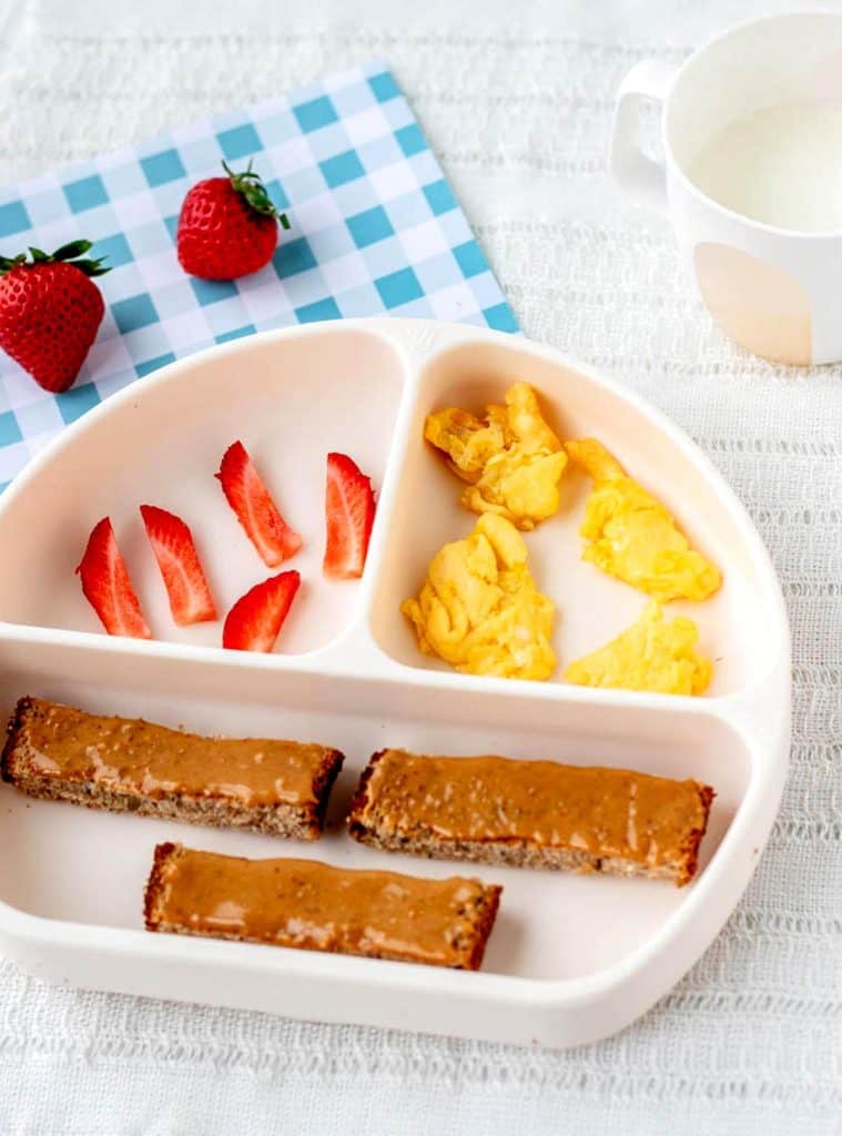 A divided plate with peanut butter toast strips, scrambled eggs and strawberries.