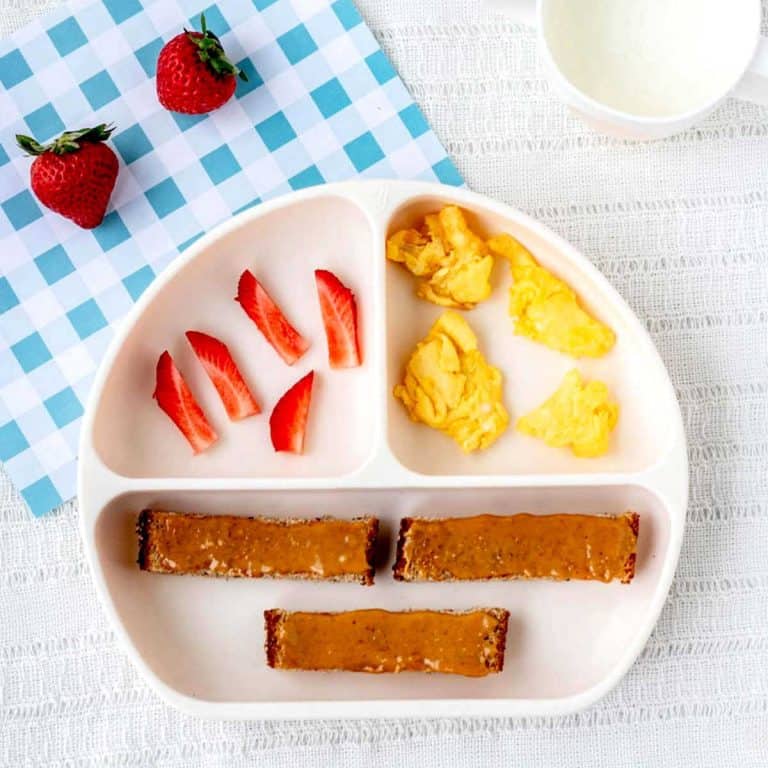 Scrambled eggs for baby on a divided plate with peanut butter toast strips and sliced strawberries.