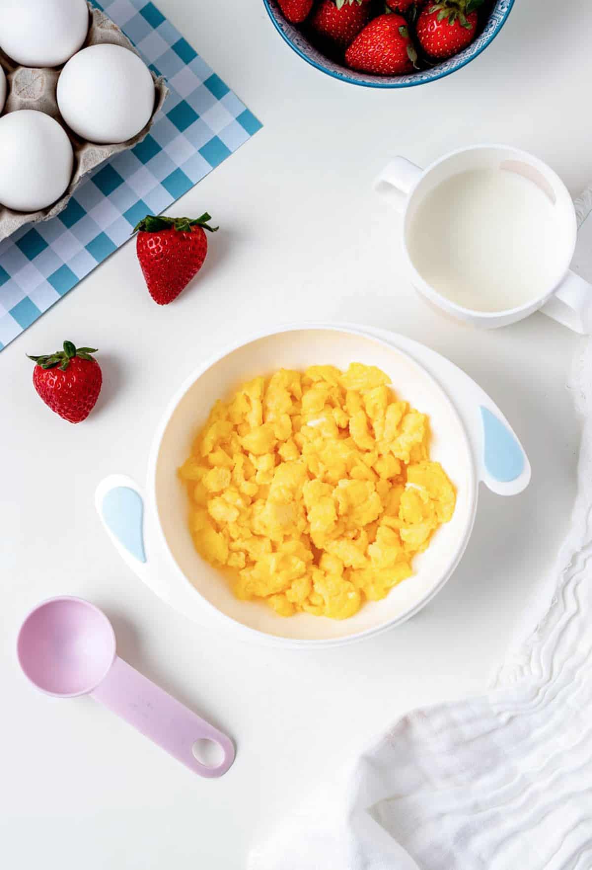 A white bowl filled with scrambled eggs for baby next to strawberries and milk.