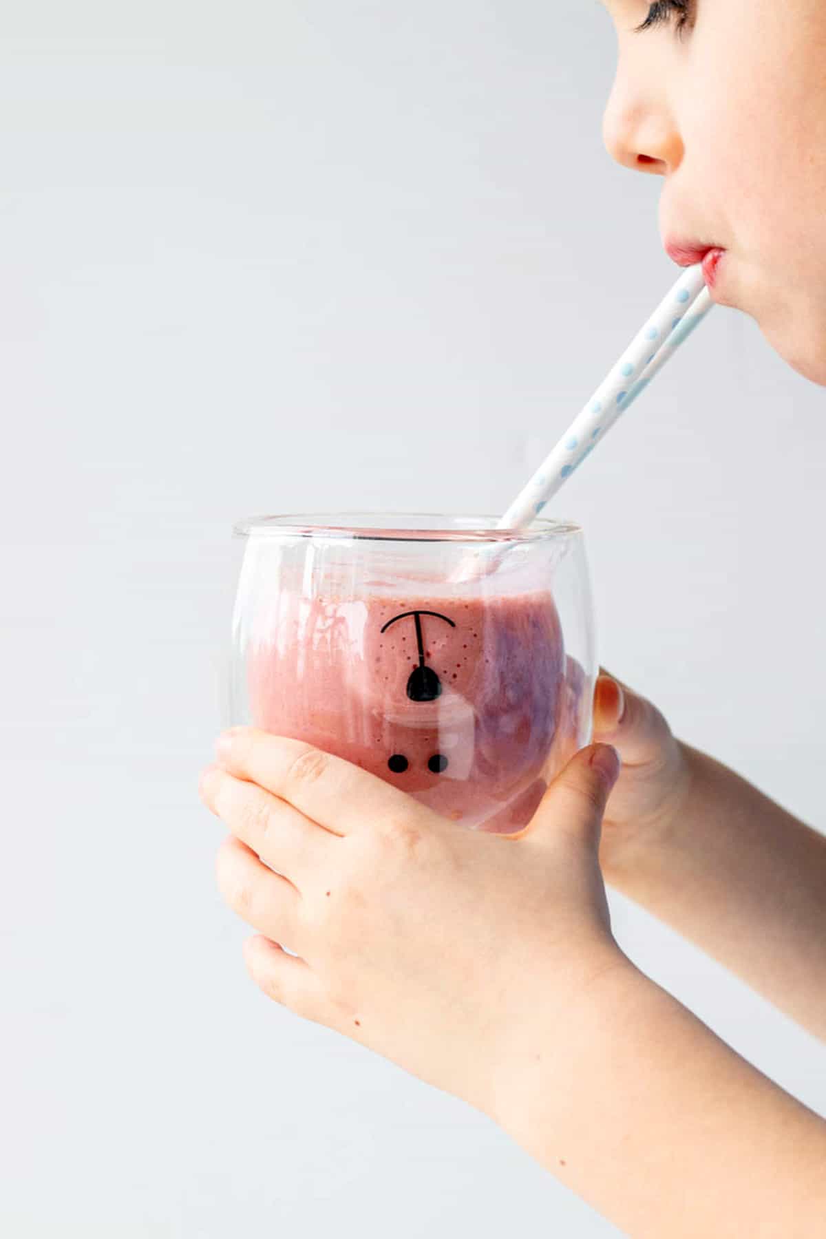 A young boy holding a glass of strawberry banana smoothie drinking some from a straw.
