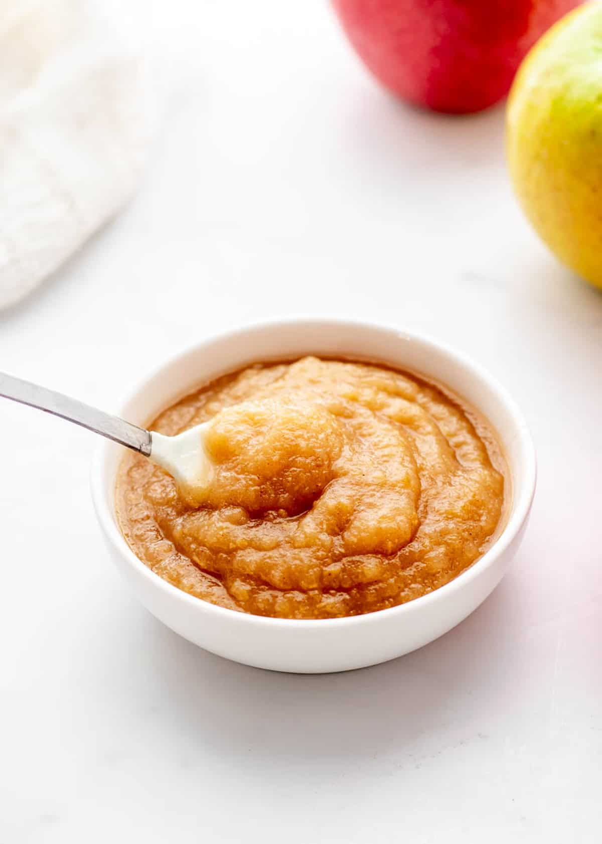 A spoon scooping up some homemade applesauce from a little white bowl.