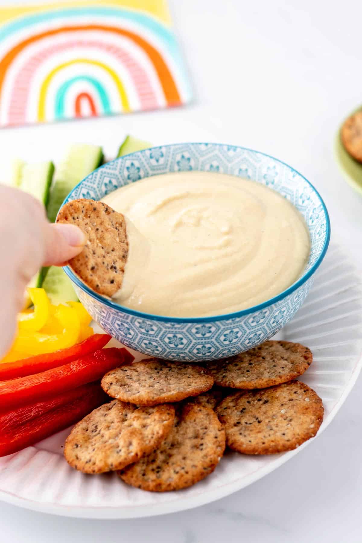 A hand dipping a pita cracker into a bowl of Greek yogurt hummus.