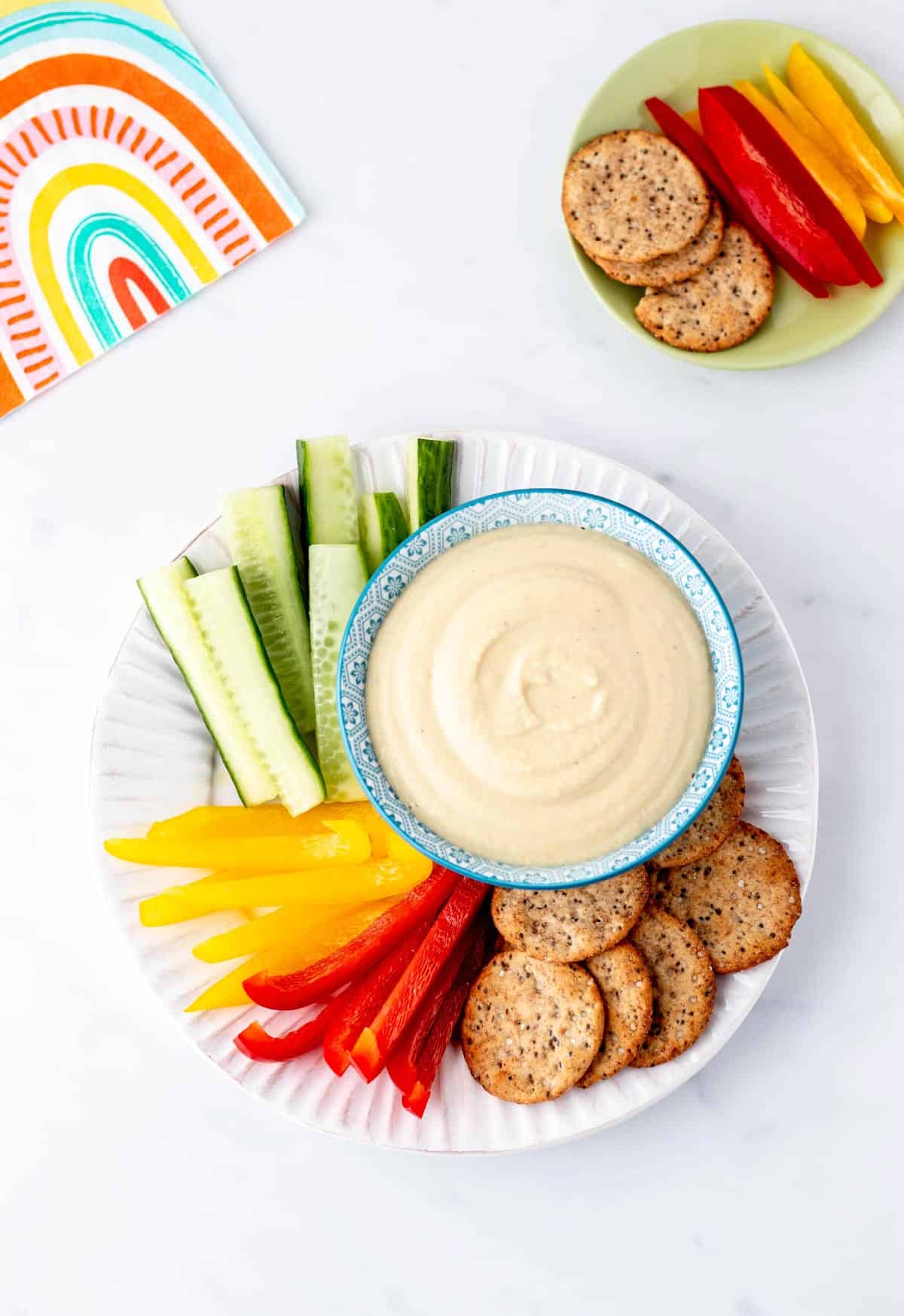 A plate with a bowl of high protein hummus and veggie sticks and crackers.