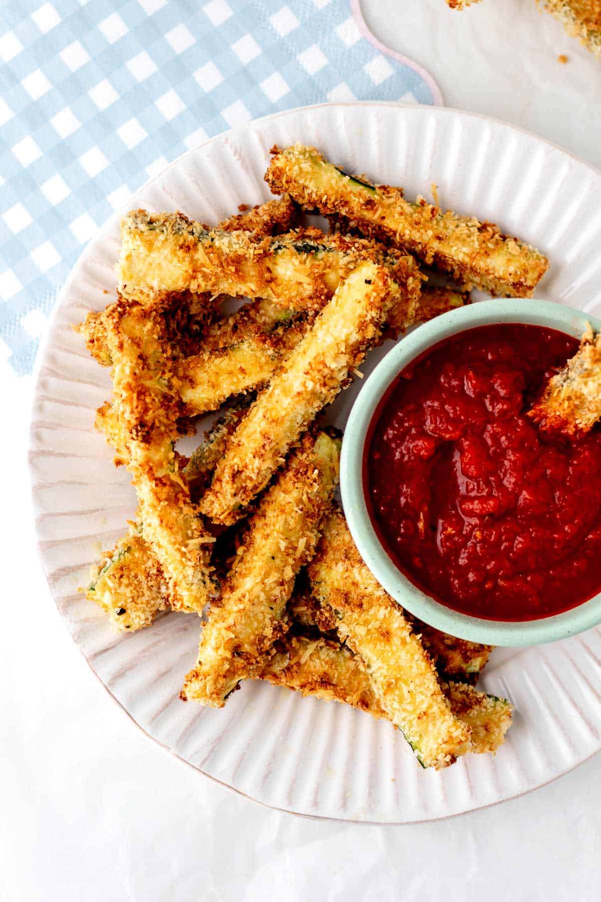 Air fryer zucchini fries on a plate with marinara next to a blue checkered napkin.