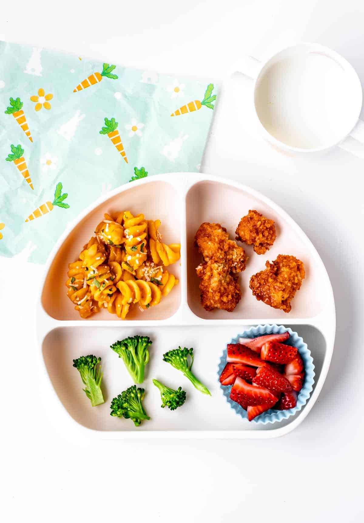 A divided plate with roasted carrot pasta, chicken bites, broccoli and strawberries.