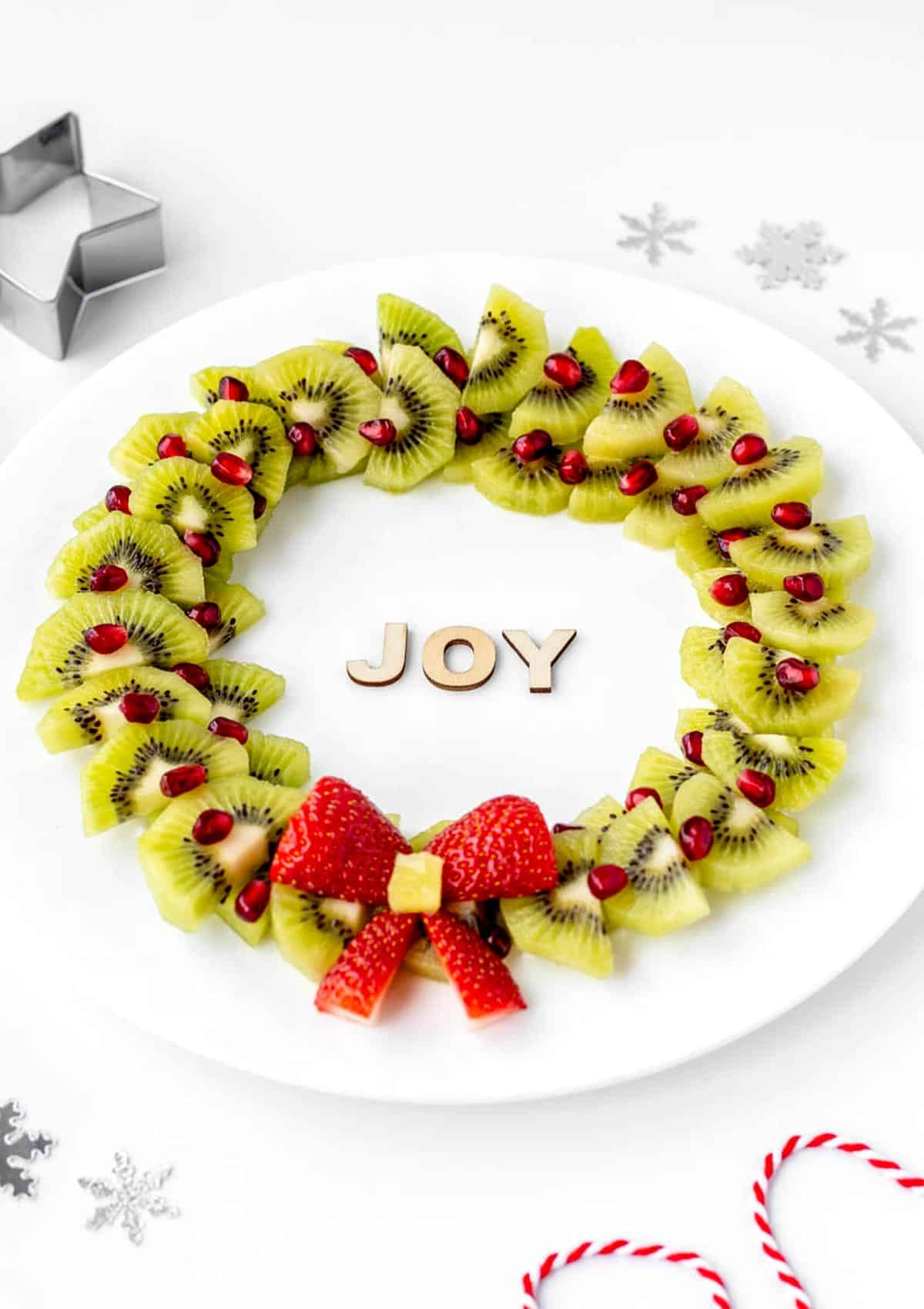 Kiwi, pomegranate and strawberry Christmas fruit wreath on a plate with the word "joy" written in the middle.