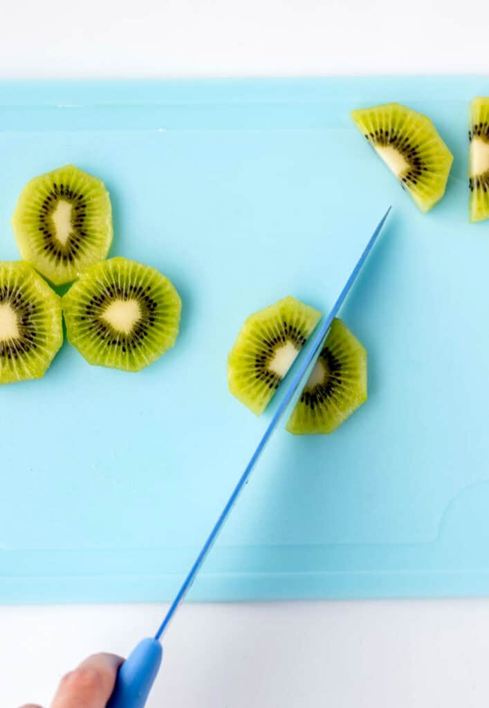 A knife slicing kiwi slices in half on a cutting board.