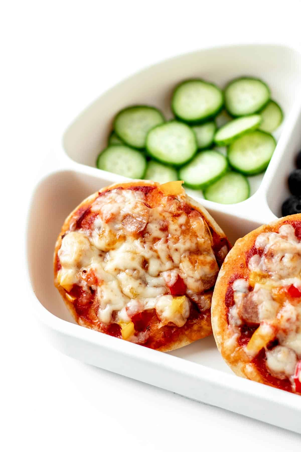 Two homemade mini pizzas on a plate with cucumbers and blueberries.