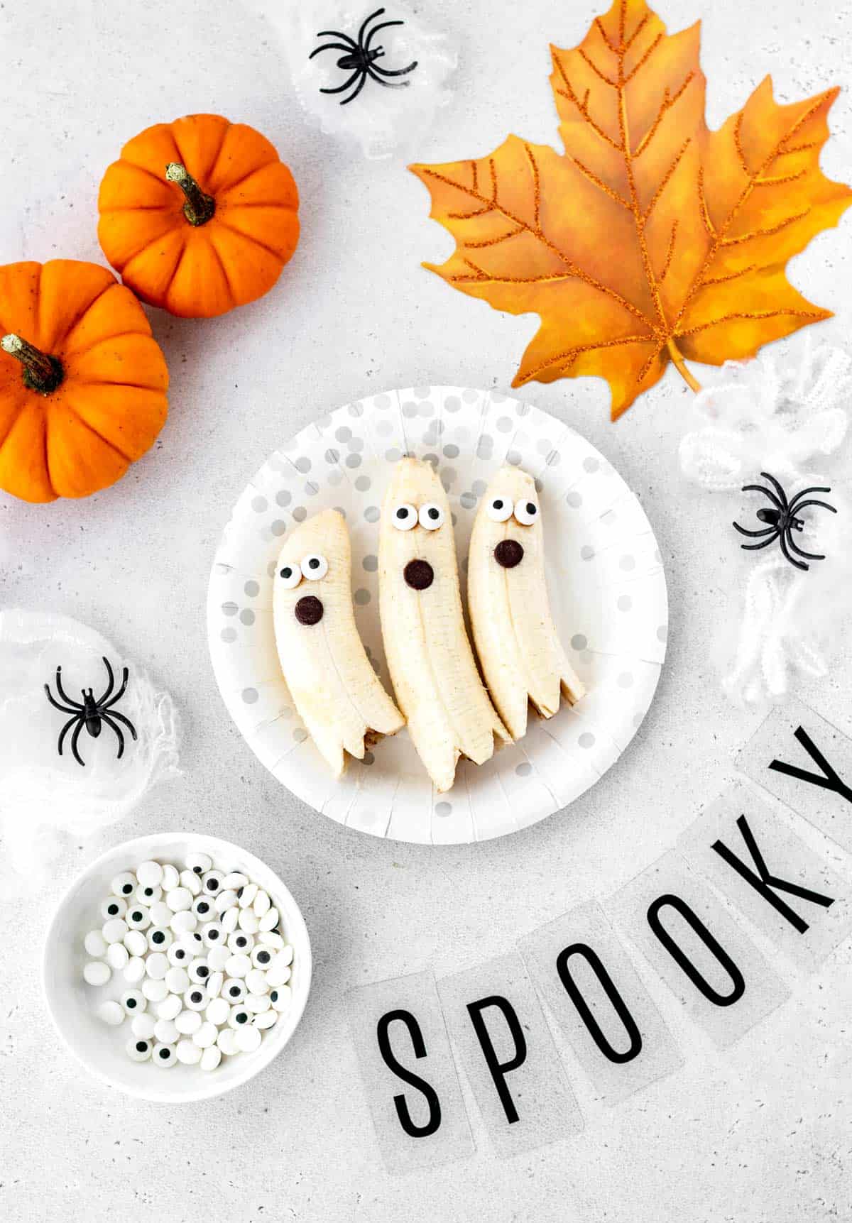 Three banana ghosts on a plate with Halloween decorations.