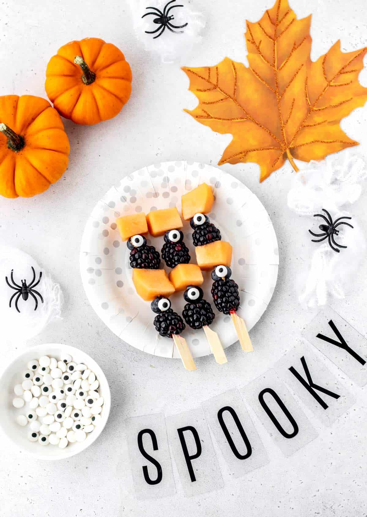 Three Halloween fruit kabobs on a plate with Halloween decorations.