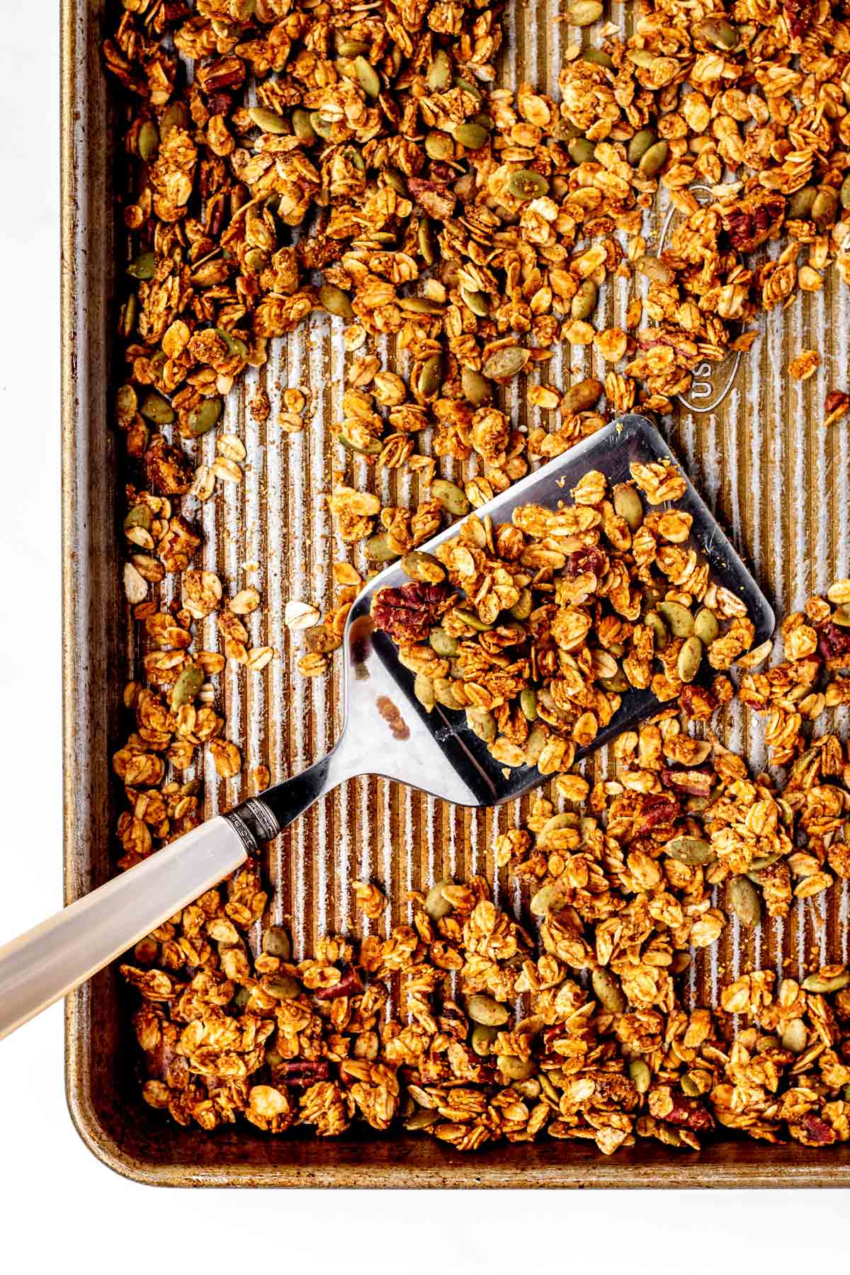 A spatula picking up some baked pumpkin seed and flax granola from a baking pan.