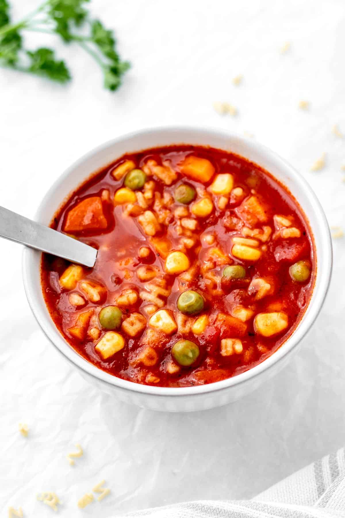 A small white bowl filled with tomato alphabet soup and a spoon.