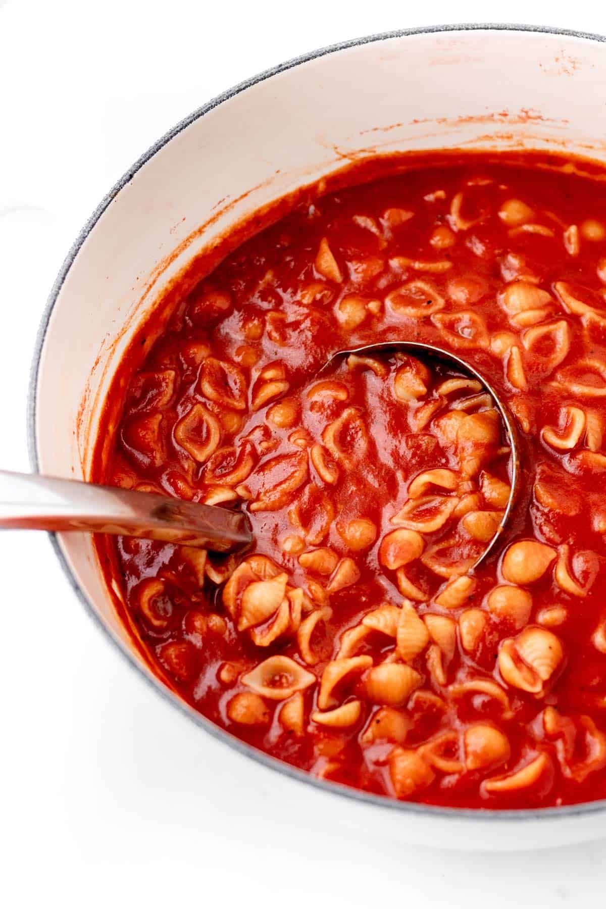 A white pot full of sea shell noodles and red sauce with a ladle.