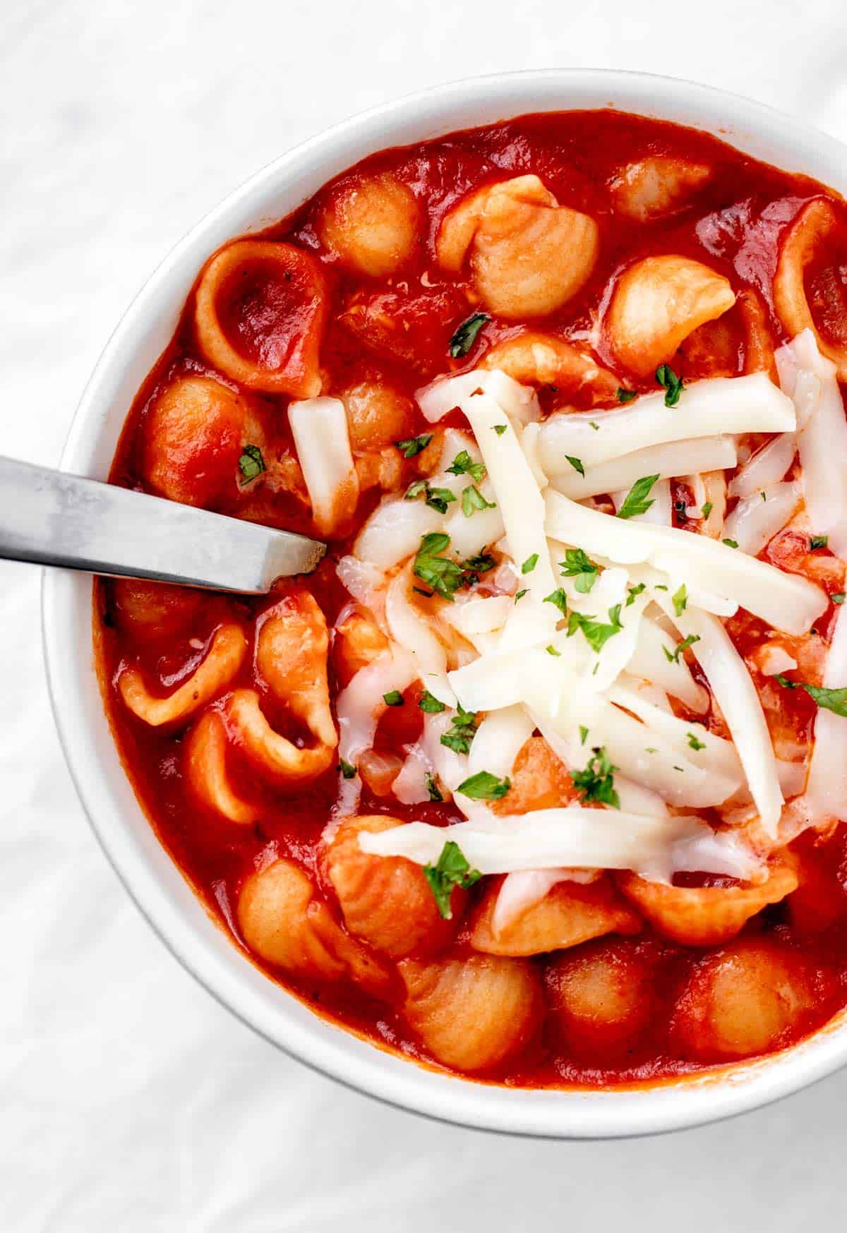 A white bowl full of noodles, red sauce and cheese on top with a spoon.