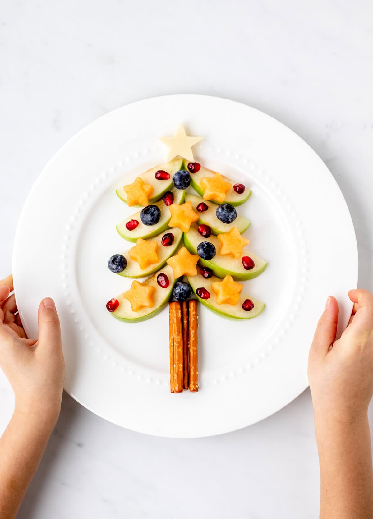 Apple Christmas tree snack on a white plate with a child's hands holding the plate.