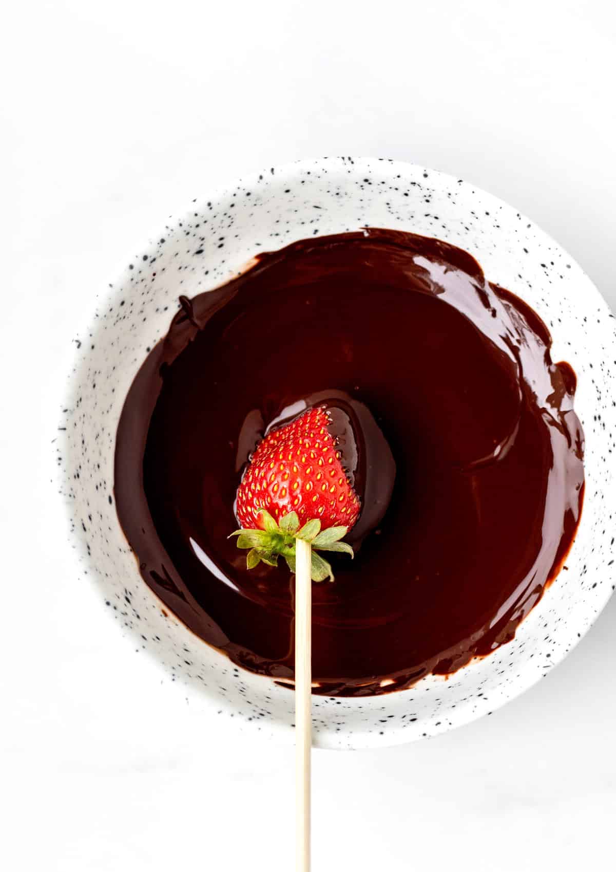 A strawberry on a wooden skewer being dipped diagonally in a bowl of melted chocolate.