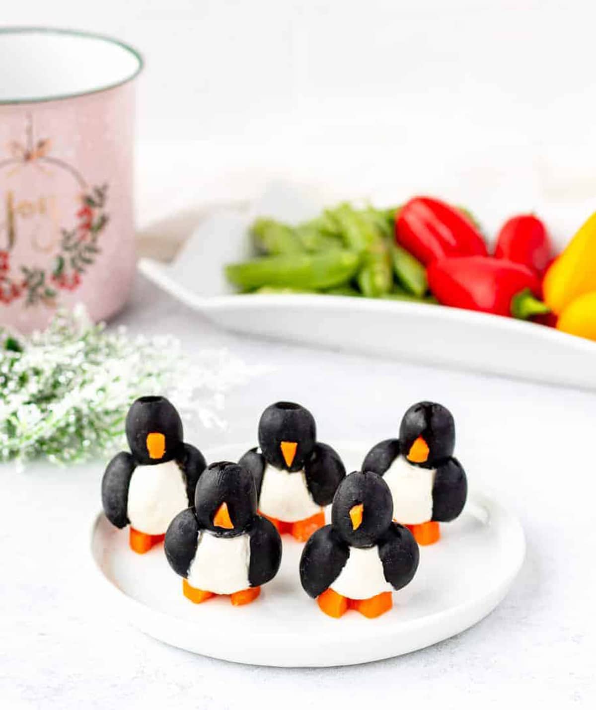 Five olive penguins on a white plate with a tray of raw vegetables in the background.