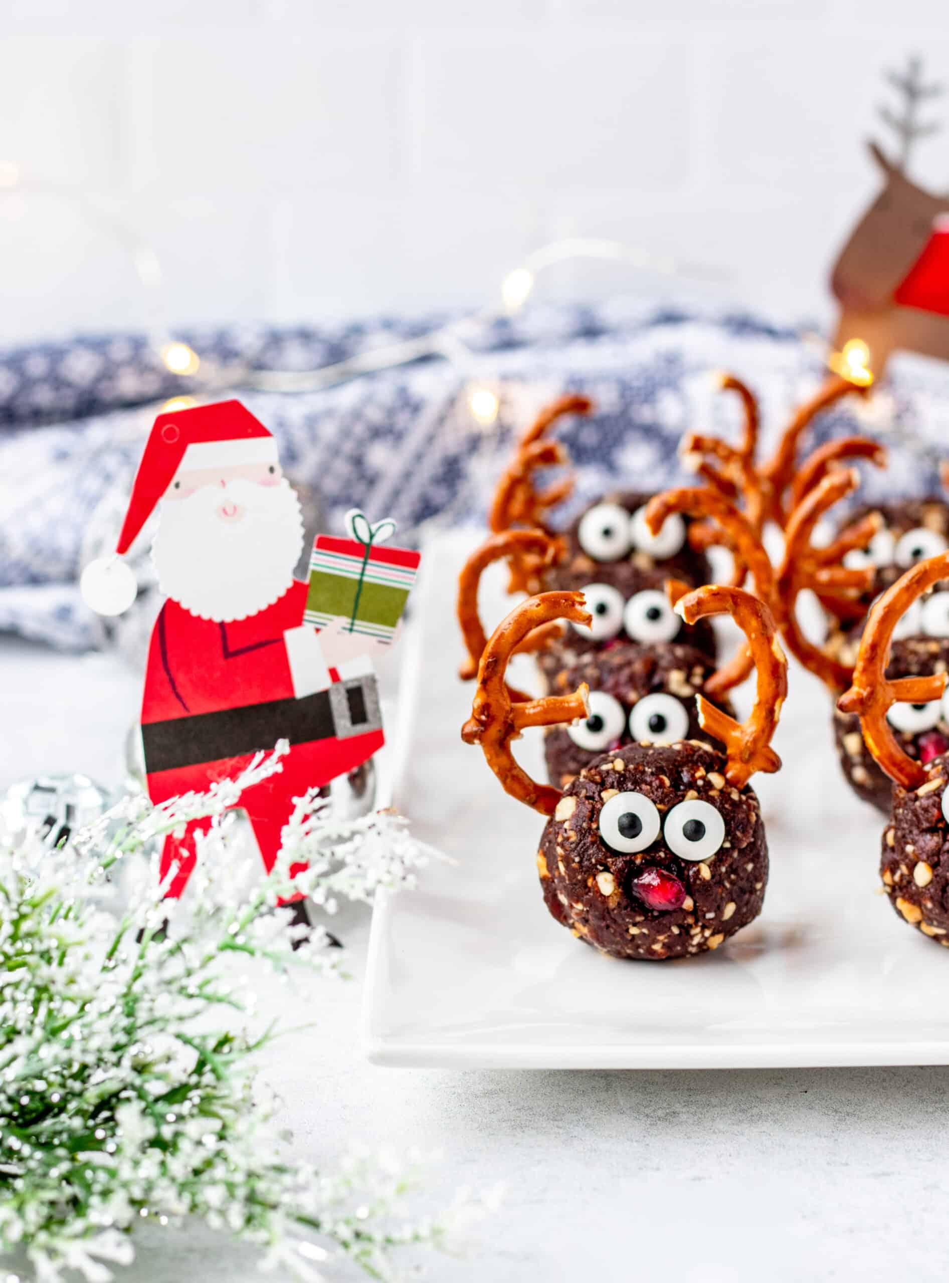 Pretzel reindeer bites lined up on a white plate, with holiday decor surrounding the plate - like a Santa Claus holding a gift.