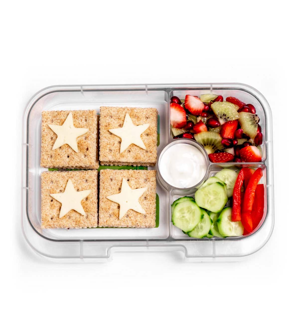 A lunch box containing whole grain crackers topped with cheese stars, a fruit medley, cucumber slices with red pepper strips and a size of tzatziki sauce.