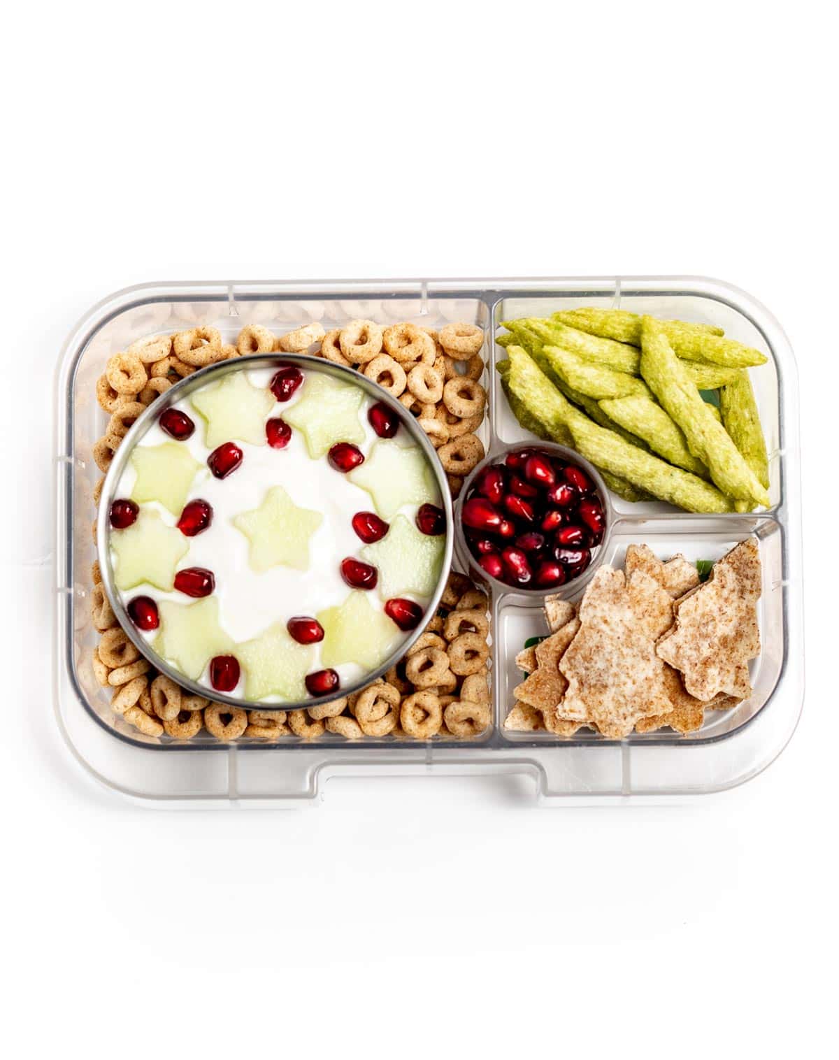 A lunchbox containing cheerios, Greek yogurt with honey melon stars and pomegranate seeds in the shape of a wreath, snap pea crisps, tortillas cut into Christmas trees, and a side of extra pomegranate seeds.