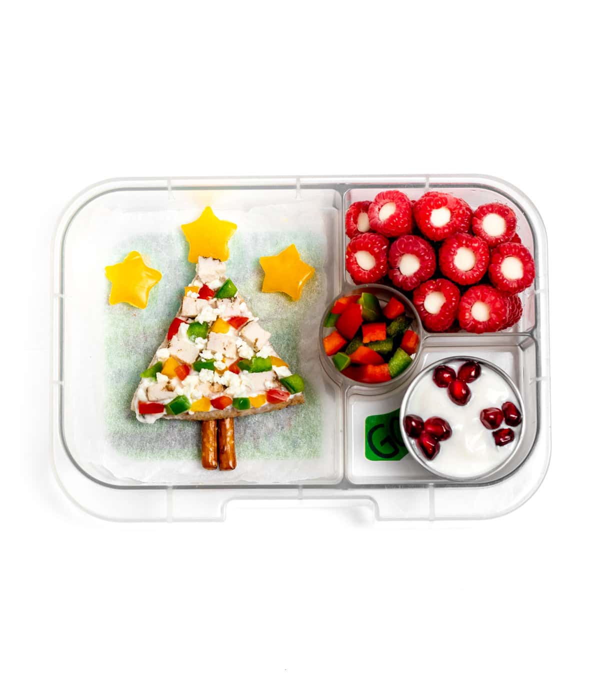 A lunchbox with a pita Christmas tree, yellow pepper mini stars, raspberries stuffed with white chocolate chips, and Greek yogurt topped with pomegrante seeds.