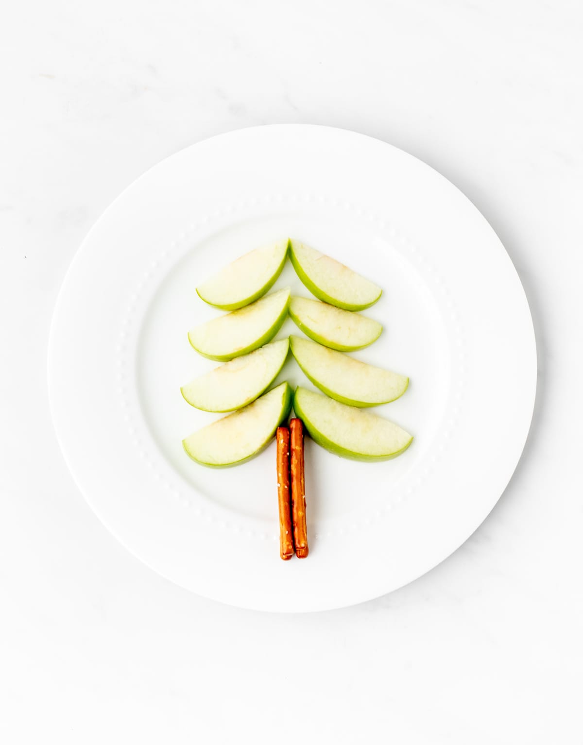 Apple slices and pretzels sticks designed to look like a Christmas tree on a white plate.