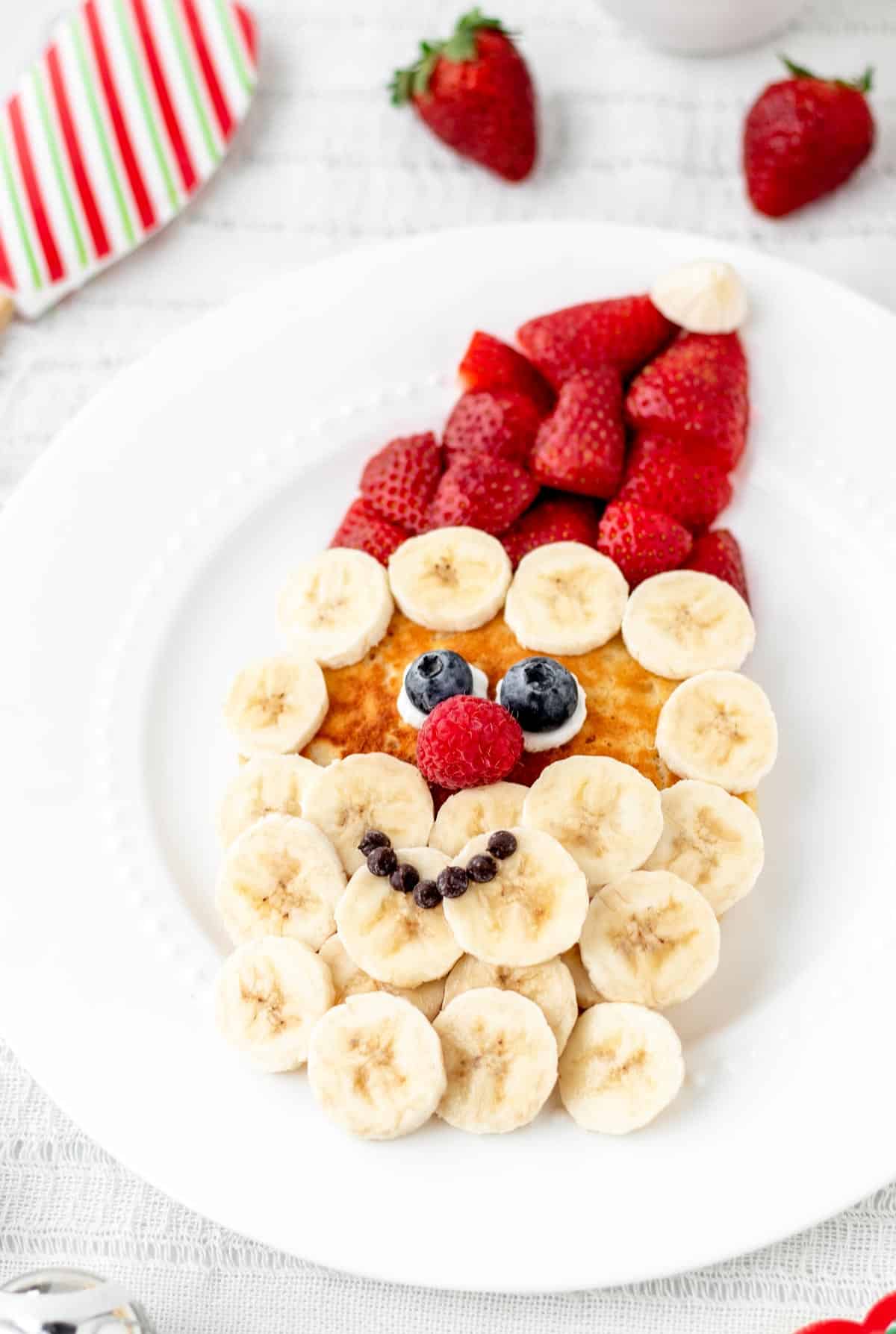 A Santa pancake with strawberries and bananas served on a large white plate.