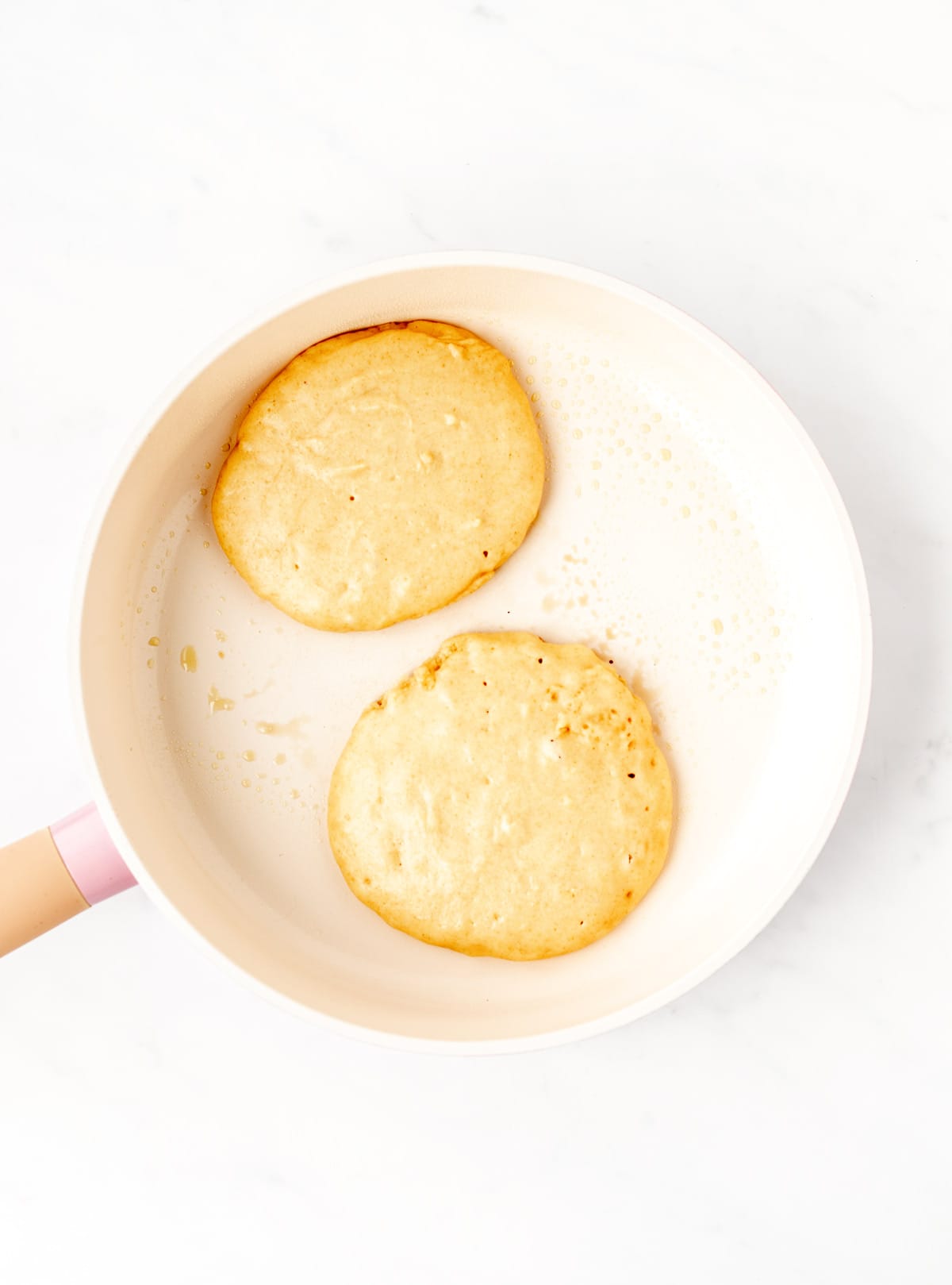 Two Greek yogurt pancakes being cooked in a frying pan.