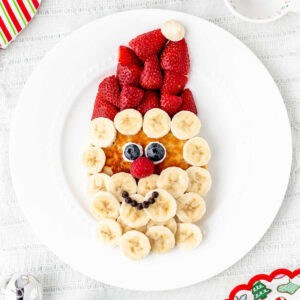 A Santa face pancake with a strawberry hat and banana beard on a white plate.