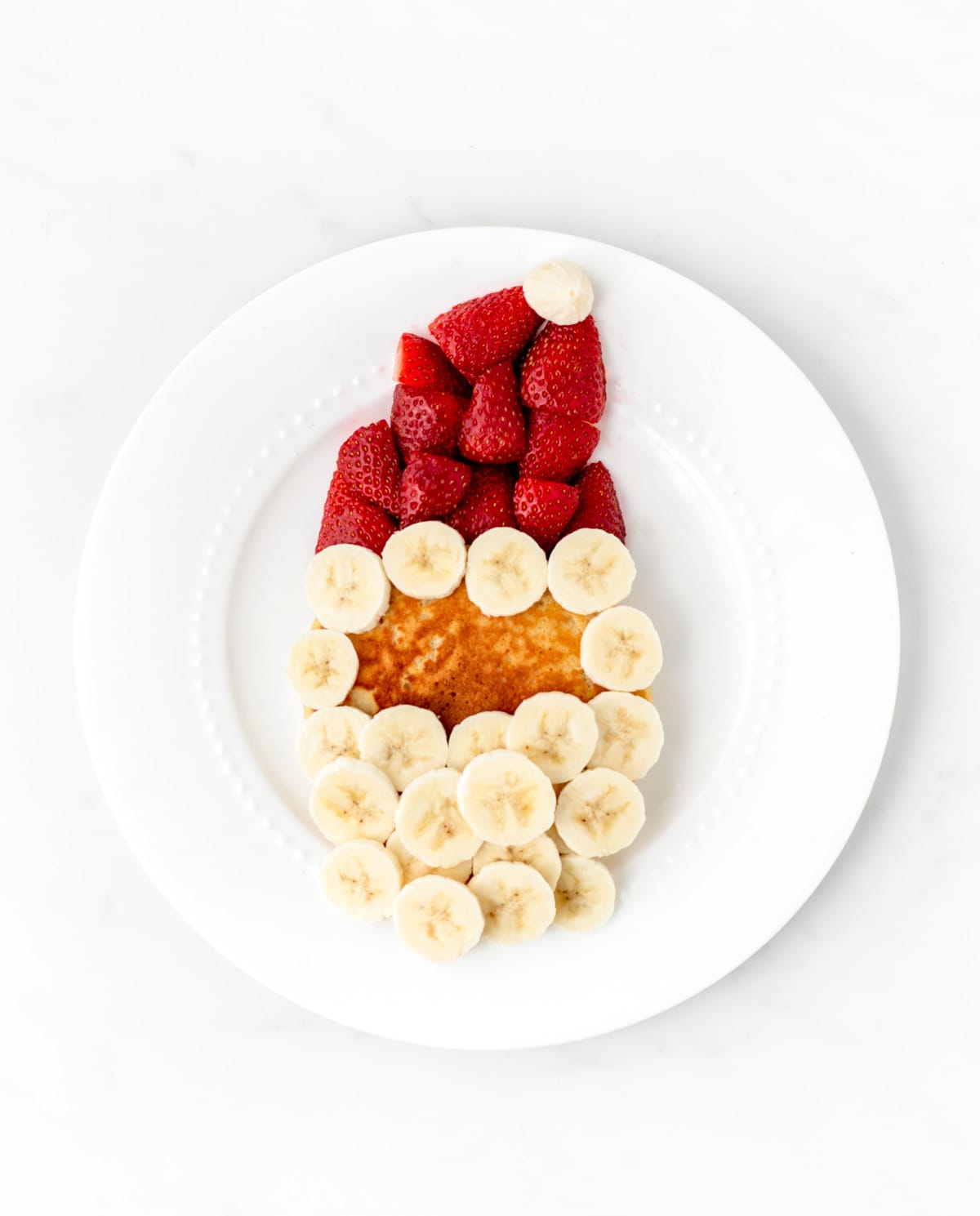 A pancake with a strawberry hat and banana beard on a plate.