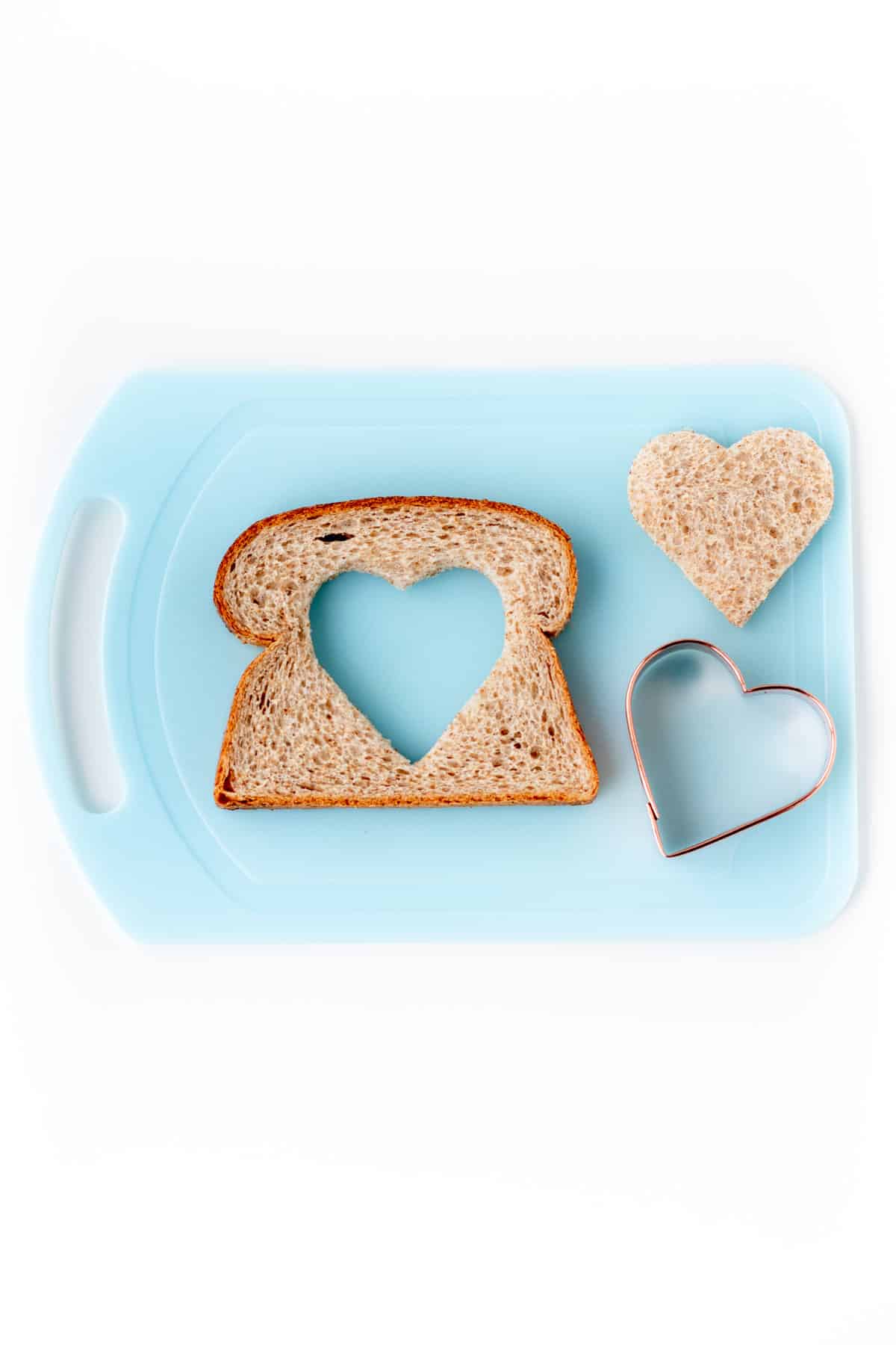 A piece of bread with a heart cut out with the heart shaped cookie cutter on a cutting board.