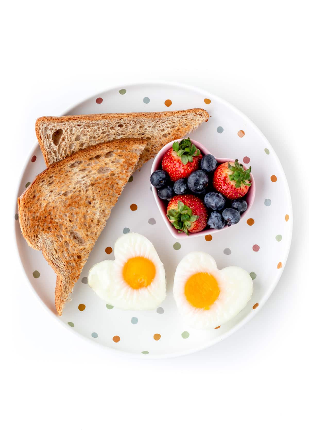 A decorative, polka dot plate with two heart shaped eggs, a sliced piece of toast, and a bowl of berries.