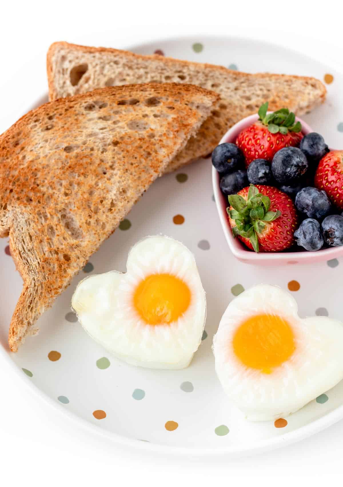 A plate of the heart shaped eggs with a piece of toast and heart-shaped bowl of mixed berries.