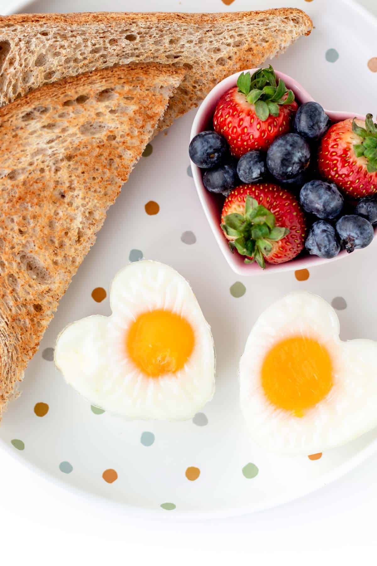 A close-up of two heart shaped eggs on a polka dot plate, alongside a sliced piece of toast and heart shaped bowl of berries.