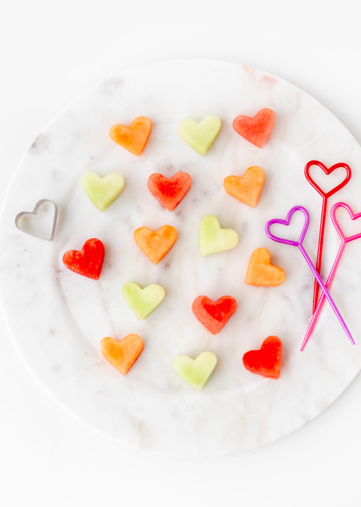 Various heart shaped fruit on a decorative plate, with the heart skewers and heart shaped cookie cutter on the side.