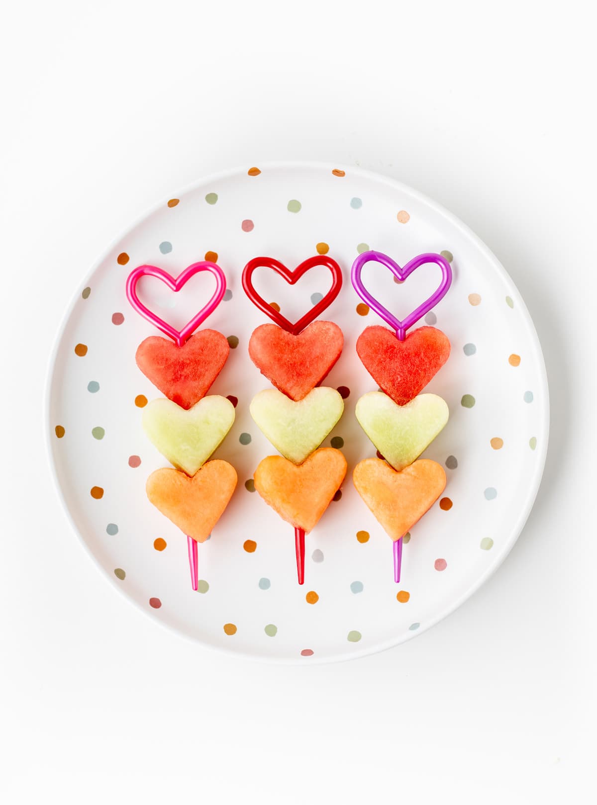 3 heart shaped fruit kabobs lined up on a decorative, polka dotted plate.