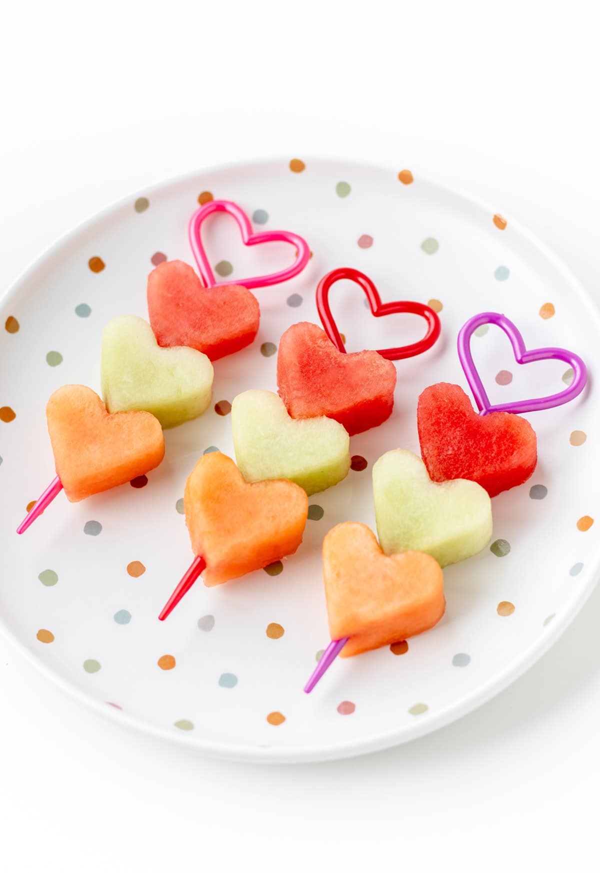 3 heart shaped fruit kabobs lined up on a polka dot plate.