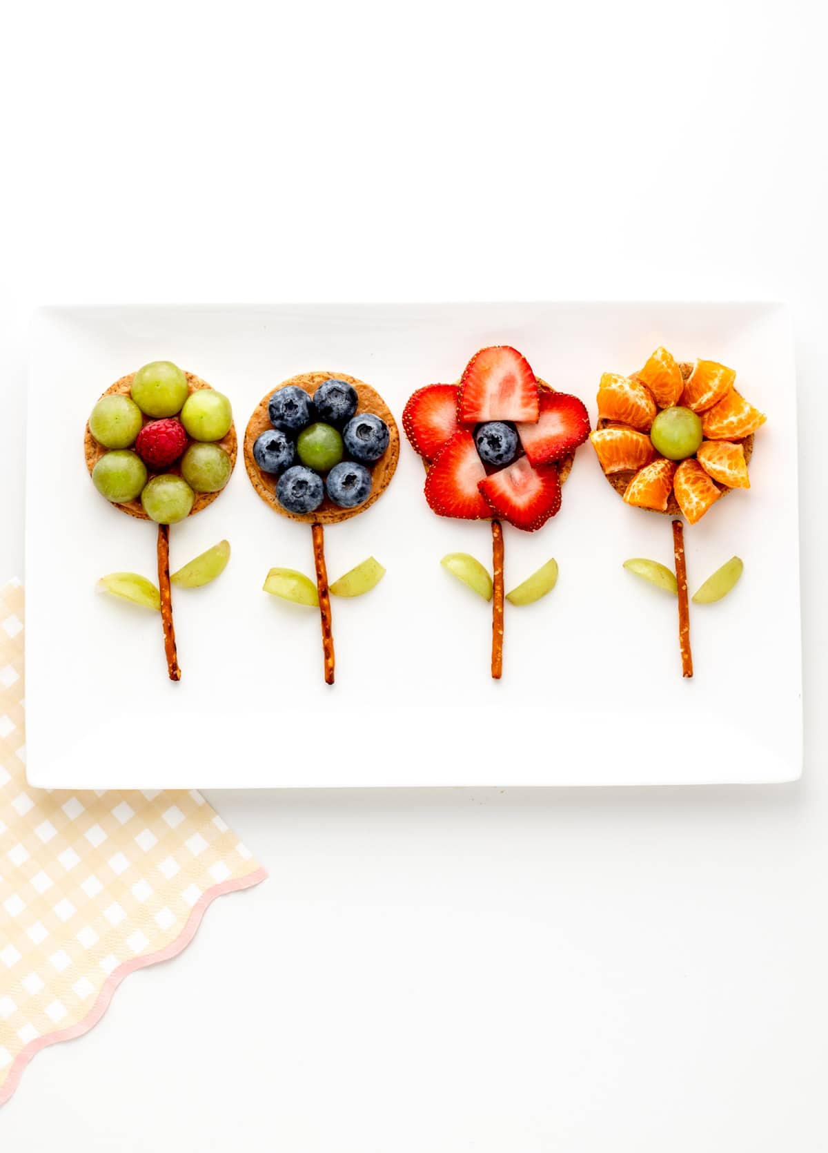 Flower crackers made of fruit and a pretzel stick stem on a white platter.