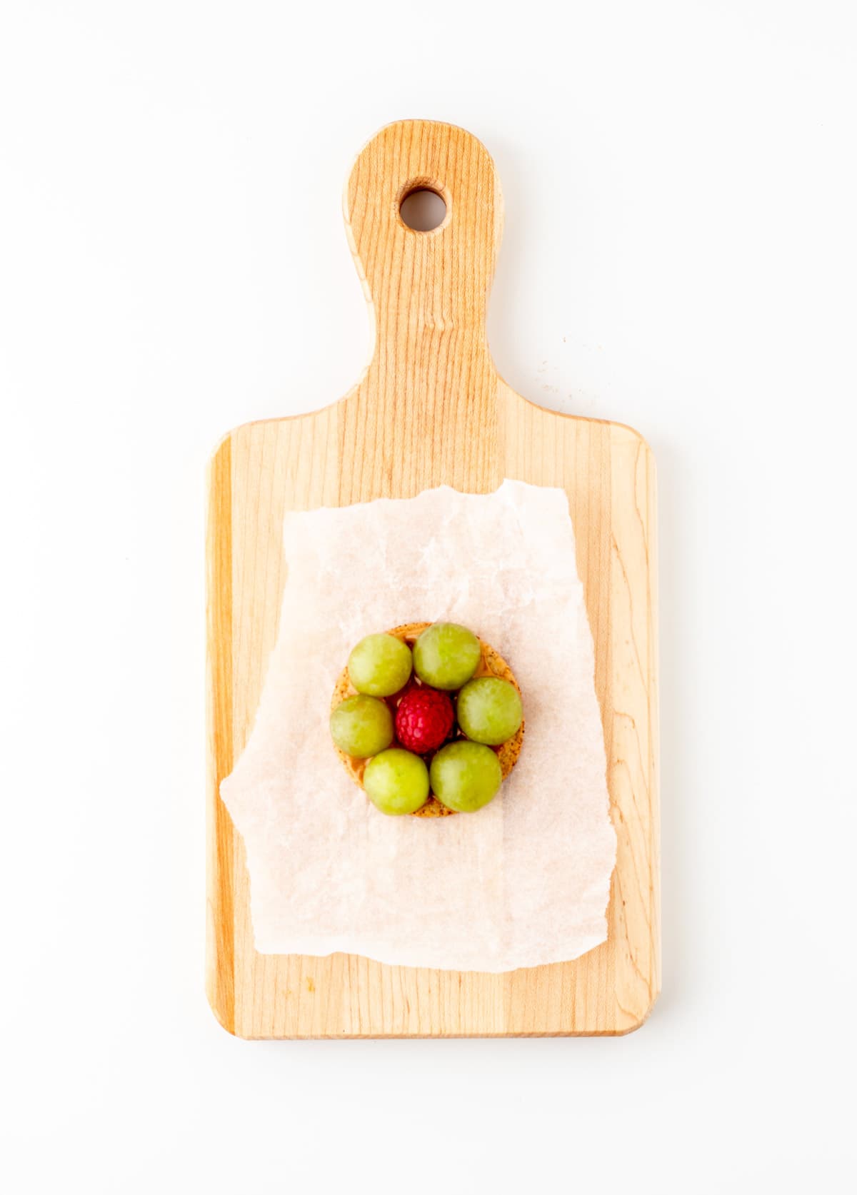 A single cracker with grapes on the perimeter with a raspberry in the center on a cutting board.