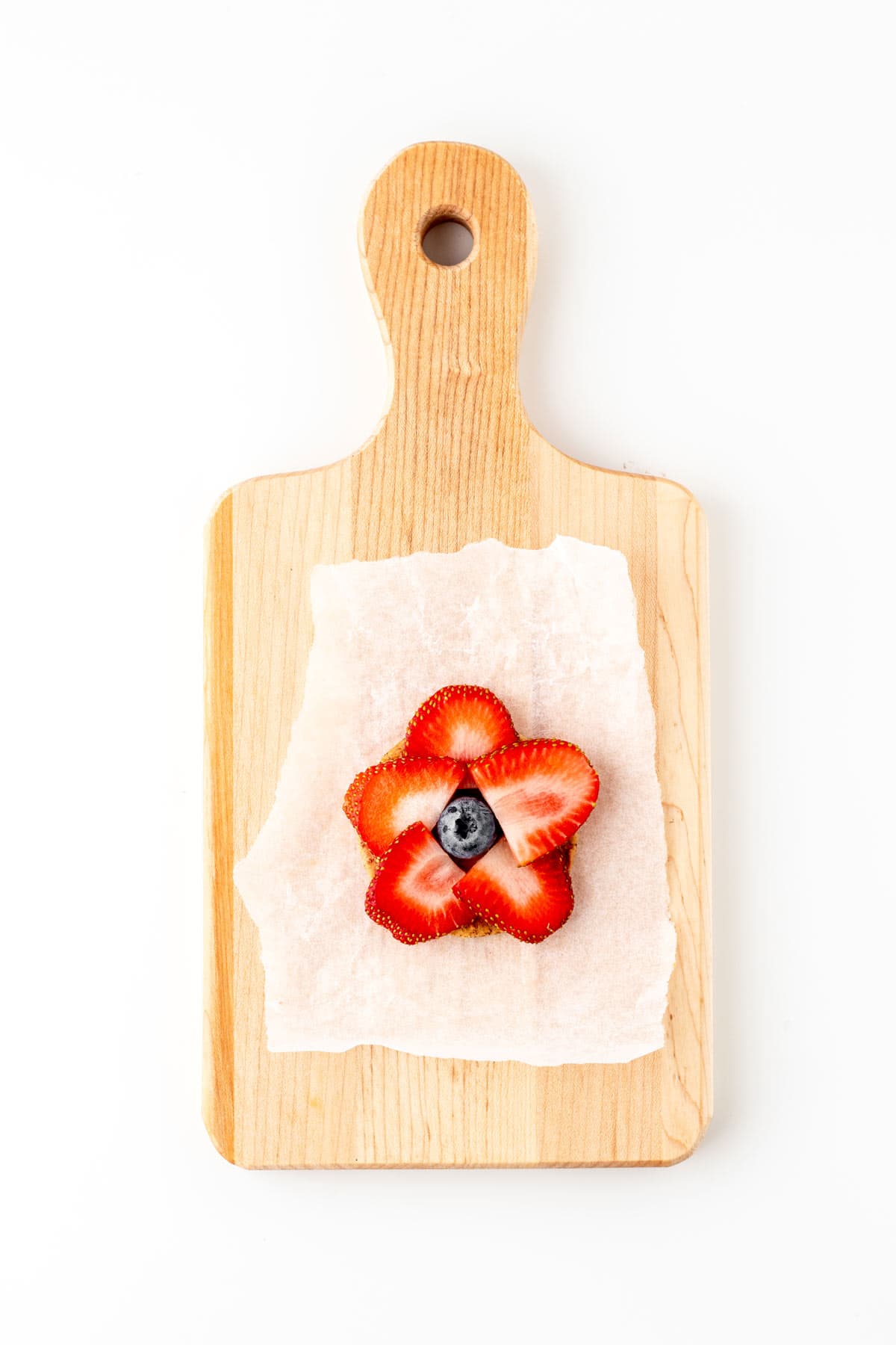 A flower cracker with cut strawberries along the perimeter with a blueberry in the center on a wood cutting board.