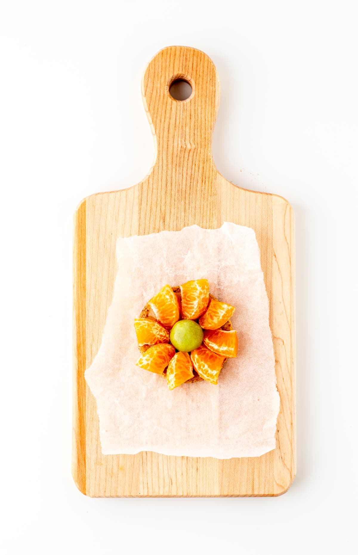 A single cracker on a wood cutting board with mandarin oranges lining the perimeter and a grape in the middle.