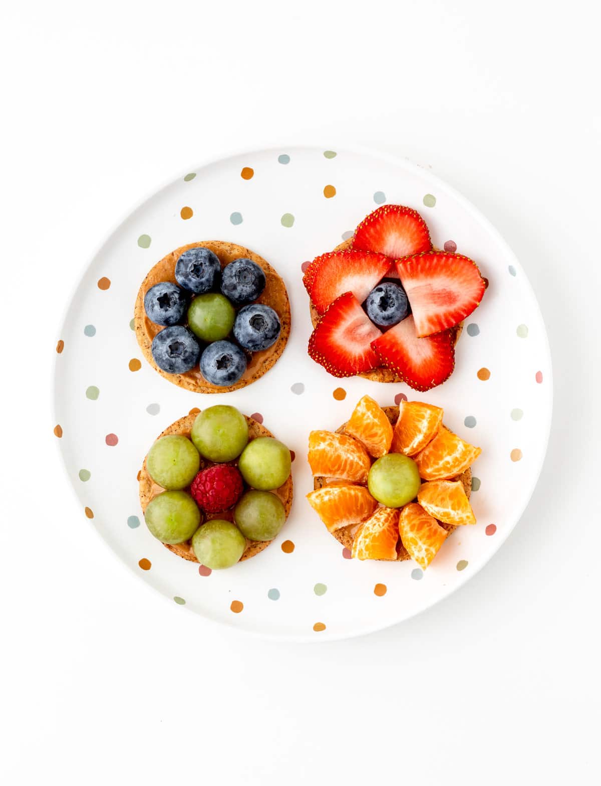 4 single crackers on a polka dotted plate: one with strawberries lining the perimeter, grapes, blueberries and then mandarin oranges.