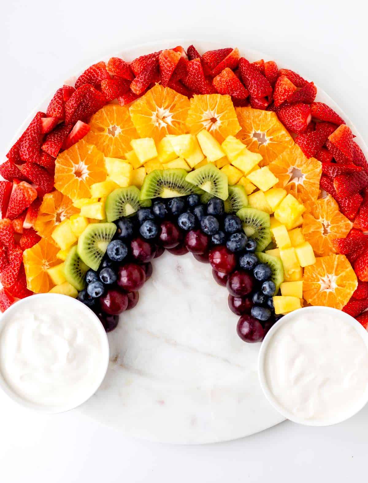 A fully assembled rainbow fruit board with two small bowls of yogurt dip on each side to resemble clouds.