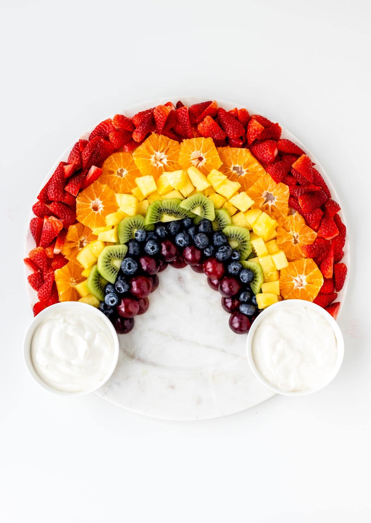 A birds-eye view of a fully assembled rainbow fruit board with two bowls of yogurt dip on each side.