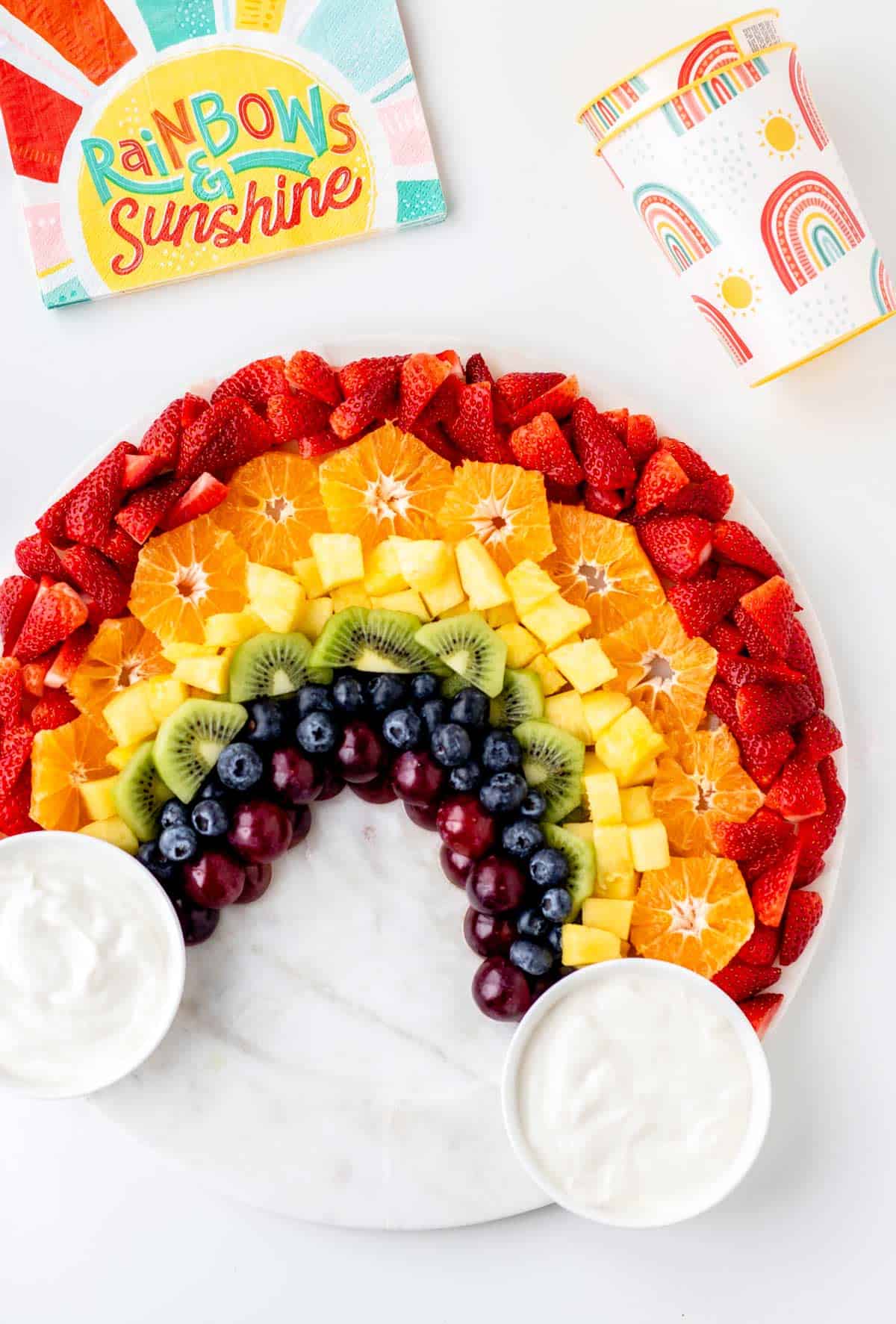 A colorful rainbow fruit tray with vanilla yogurt dip in small bowls at each side of the rainbow to resemble clouds.