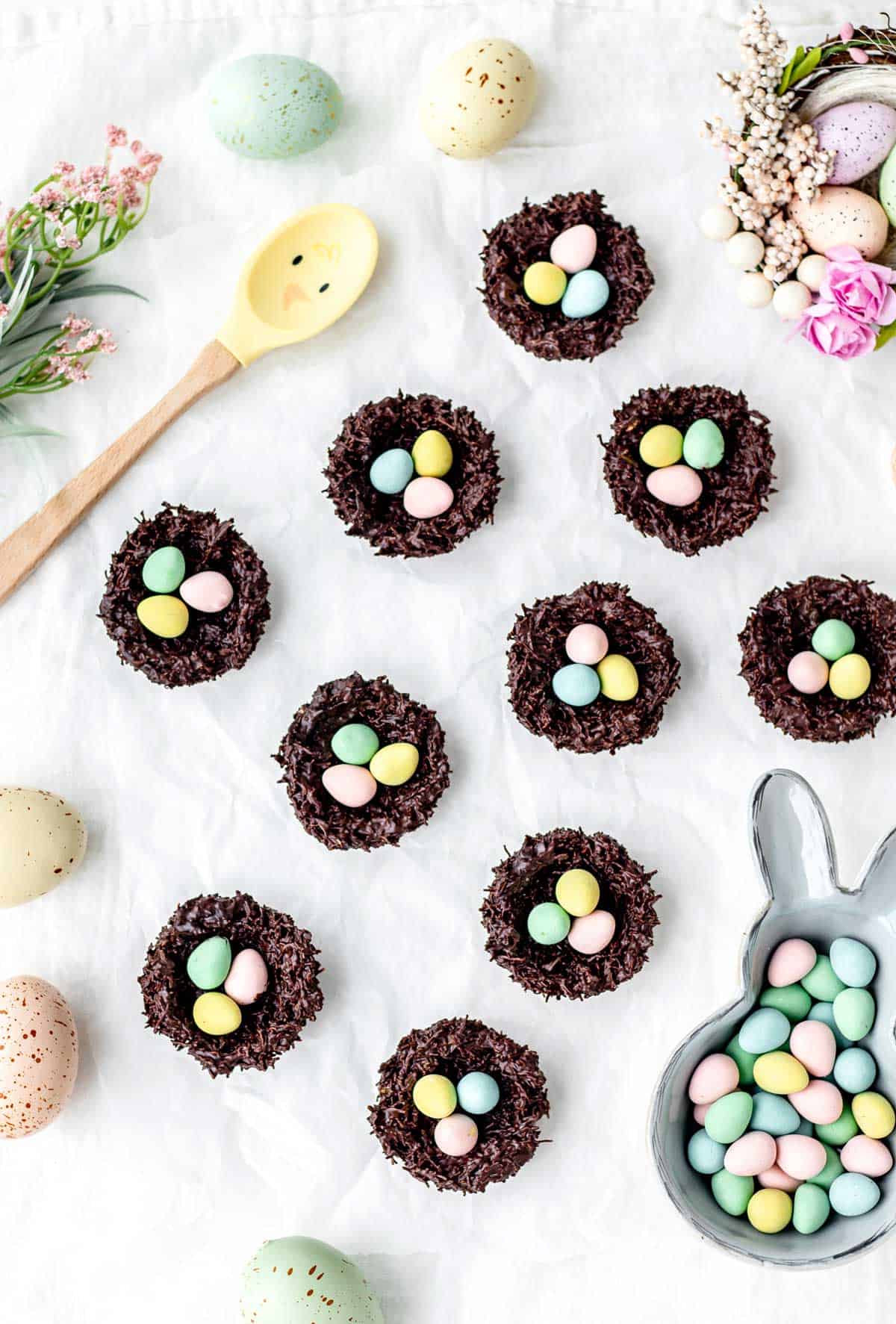 Shredded wheat Easter nests spaced out on a countertop with along with a spoon and a bowl of mini chocolate eggs.