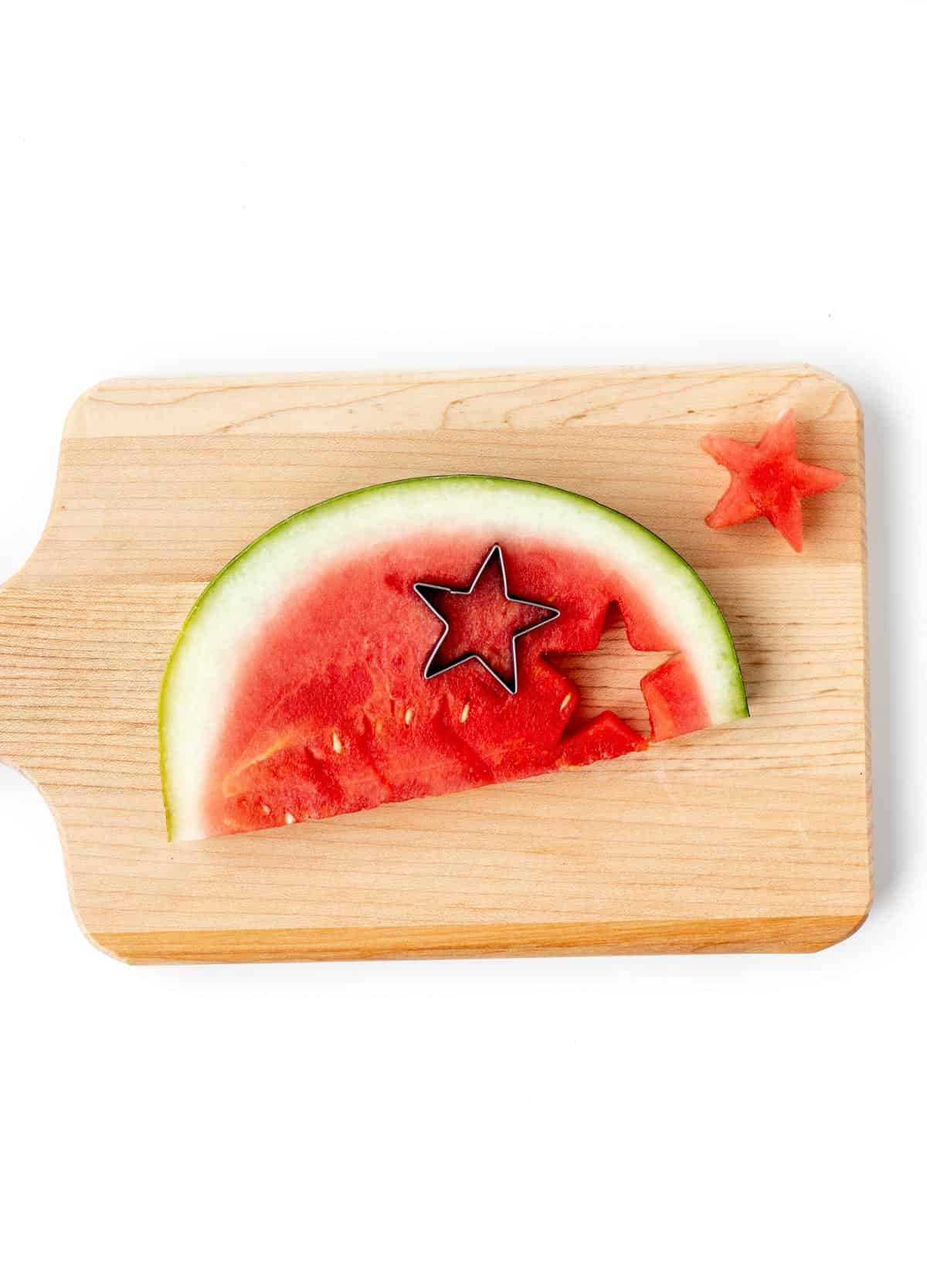 A star cookie cutter making star shapes out of a watermelon slice on a wooden cutting board.
